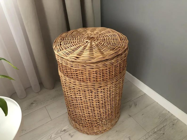 May include: A tall, cylindrical wicker laundry hamper with a domed lid. The hamper is made of woven light brown wicker and sits on a light-colored wooden floor. The lid has a central knob.