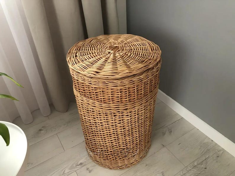 May include: A cylindrical wicker laundry hamper with a domed lid. The hamper is made of woven light brown wicker and sits on a light-colored wooden floor. The lid has a central knob.