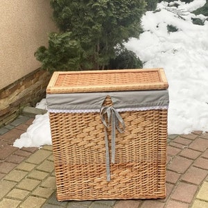 May include: A woven wicker laundry basket with a gray fabric lid and a gray ribbon. The basket is sitting on a brick patio.