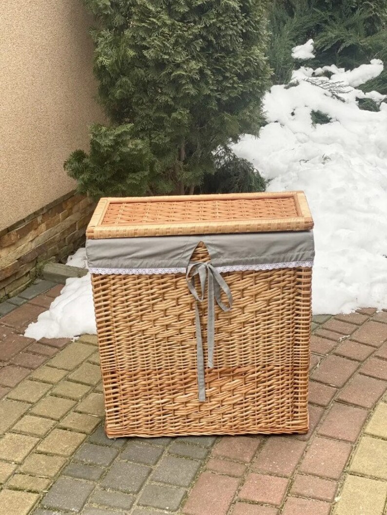 May include: A woven wicker laundry basket with a gray fabric lid and a gray ribbon. The basket is sitting on a brick patio.