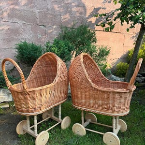 Puede incluir: Dos cochecitos de muñecas antiguos de mimbre. Cada cochecito tiene un cuerpo de cesta tejida, un asa curva y está montado sobre un marco de madera con cuatro ruedas. Los cochecitos son de color marrón claro y están sobre una superficie herbosa.