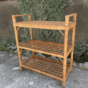 Può includere: Una mensola in vimini marrone chiaro a tre livelli di forma rettangolare. La mensola presenta un motivo intrecciato sui ripiani e sui lati. Il ripiano superiore ha una maniglia. La mensola è realizzata con materiali naturali ed è adatta allo stoccaggio.