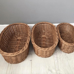 Op de afbeelding: Drie bruine rieten manden in verschillende maten. De ovale manden hebben een geweven ontwerp. De grootste mand staat links, de middelste in het midden en de kleinste rechts. De manden zijn leeg en staan op een licht houten vloer.