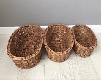 May include: Three brown wicker baskets of varying sizes. The baskets are oval-shaped with a woven design. The largest basket is on the left, the medium in the center, and the smallest on the right. The baskets are empty and sit on a light-colored wooden floor.