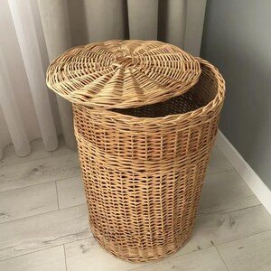May include: A natural woven wicker laundry hamper with a matching lid. The hamper is cylindrical with a slightly flared top and a lid that is partially open. The wicker has a light brown color and a textured surface.