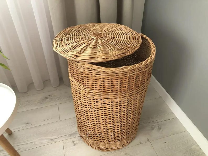 May include: A natural woven wicker laundry hamper with a matching lid. The hamper is cylindrical with a slightly flared top and a lid that is partially open. The wicker has a light brown color and a textured surface.