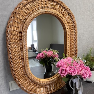 May include: Oval-shaped wicker mirror with a natural brown finish. The mirror is surrounded by a thick, woven frame. The mirror is reflecting a bouquet of pink roses in a black vase.