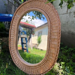 May include: Oval-shaped wicker mirror with a natural brown finish. The mirror has a woven frame and a clear glass reflection.