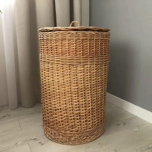 May include: A tall, cylindrical wicker laundry hamper with a matching lid. The hamper is woven from light brown wicker and has a rounded base. The lid has a small handle. The hamper is placed in a room with a neutral color scheme.