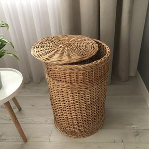 May include: A natural woven wicker laundry hamper with a removable lid. The hamper is cylindrical with a woven pattern and a lid that is slightly ajar. The hamper is a light brown color and is in a room with a white table and a plant.