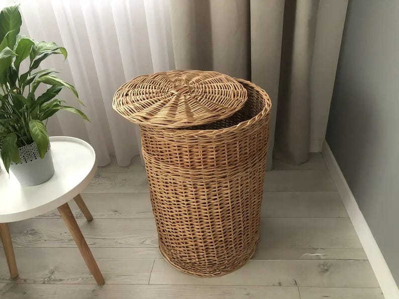 May include: A natural woven wicker laundry hamper with a removable lid. The hamper is cylindrical with a woven pattern and a lid that is slightly ajar. The hamper is a light brown color and is in a room with a white table and a plant.