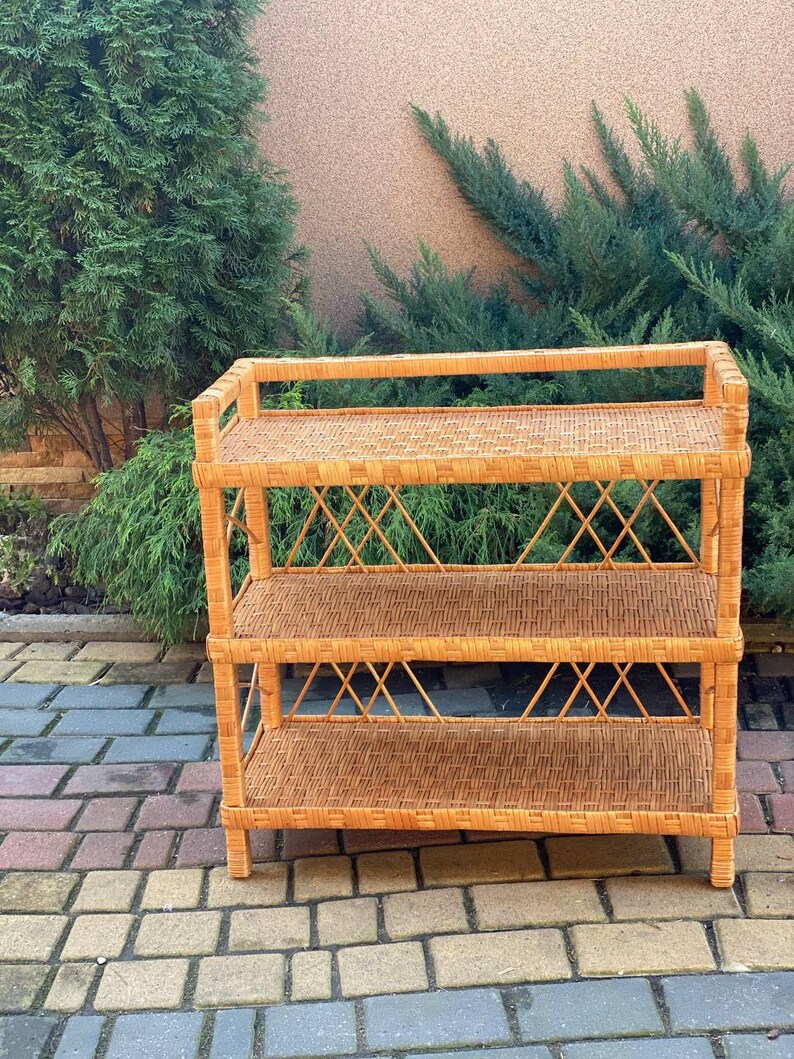 May include: A three-tiered wicker shelf unit with a natural brown finish. The shelf is made of woven rattan and has a simple, rustic design.