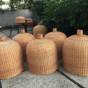 Könnte beinhalten: Eine Sammlung von gewebten Rattan-Lampenschirmen in verschiedenen Größen. Die Lampenschirme haben eine abgerundete, glockenförmige Form mit einem zylindrischen Hals. Die natürliche, hellbraune Farbe des Rattans erzeugt eine warme Ästhetik. Geeignet für Innen- und Außenbereiche.