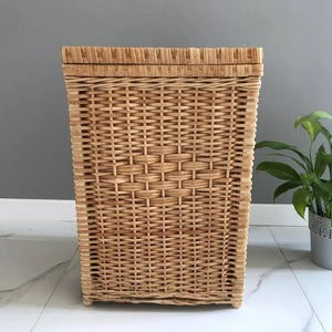 May include: A rectangular, light brown wicker laundry hamper with a lid. The hamper is made of woven wicker and has a textured surface. The hamper is designed to store laundry and is a decorative storage solution.