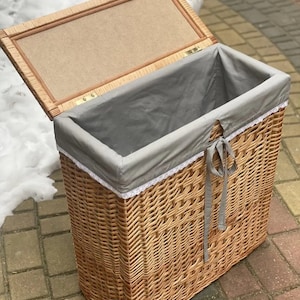 May include: A wicker laundry basket with a gray fabric liner and a white lace trim. The lid is open and the basket is sitting on a brick patio.