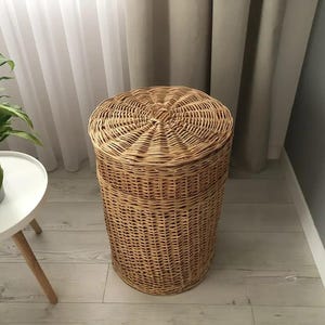 May include: A cylindrical, woven wicker basket with a lid. The basket is light brown and stands on a light-colored wooden floor. The lid is round and fits snugly on top. The basket is next to a small white table with a plant.