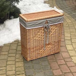 May include: A woven wicker laundry basket with a gray fabric lid and white lace trim. The basket is sitting on a brick patio.