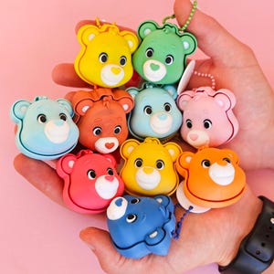 May include: A collection of nine small, colourful silicone bear-shaped charms, each featuring a distinct facial expression and a small attached ball chain. The bears are rendered in shades of yellow, green, light blue, orange, pink, red, and dark blue.