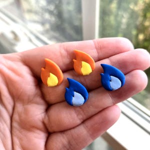 Puede incluir: Cuatro pendientes en forma de llama, en naranja, amarillo y azul. Los pendientes son pequeños, con un diseño detallado. Dos son naranjas con centros amarillos, y dos son azules con centros azul claro. Se sostienen en una mano.