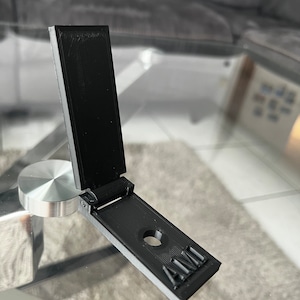 Op de afbeelding: Een zwarte 3D-geprinte telefoonstandaard met een scharnier en een rond gat. De standaard is ingeklapt en het woord "AMD" is op de onderkant gedrukt.