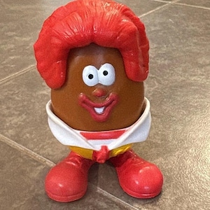 May include: A brown Mr. Potato Head toy with red hair, white eyes, and a red smile. It wears a white collar with a yellow and red outfit and red shoes. A fun, collectible toy.