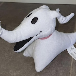 Puede incluir: Peluche blanco de Zero, el perro fantasma de Pesadilla antes de Navidad. El juguete tiene ojos negros, boca negra y un collar rosa. La nariz del perro es una calabaza. Una etiqueta está adjunta al juguete.