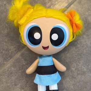 May include: A doll of a cartoon character with yellow hair styled in pigtails with orange and yellow bows. The doll has large blue eyes, a light blue dress with a black stripe, white socks, and black shoes.