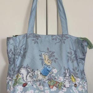 May include: A light blue tote bag featuring a printed pattern of Peter Rabbit characters, including a squirrel, a butterfly, and a bee. The design includes floral elements, leafy branches, and a zipper closure at the top with two fabric handles.