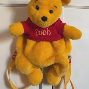 May include: A plush Winnie the Pooh backpack featuring a bright yellow bear with a soft texture, wearing a red shirt with the name Pooh embroidered in gold thread. The backpack includes adjustable yellow fabric straps and a small blue tag on the side.