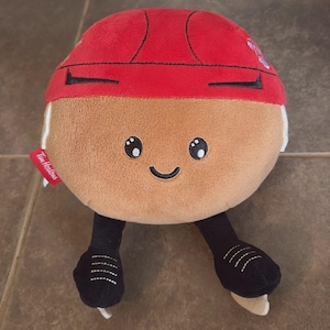 May include: A round, tan plush toy shaped like a Tim Hortons Timbits doughnut hole, wearing a red hockey helmet and black ice skates. The plush features embroidered black eyes and a smile, with a small red tag on the side displaying the Tim Hortons logo.