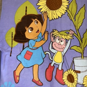 Puede incluir: Una manta de forro polar morada con una imagen de dibujos animados de Dora la Exploradora y Botas el mono. Dora está alcanzando un girasol amarillo y Botas está sosteniendo un girasol. La manta tiene la palabra "Dora" impresa en amarillo en la parte inferior.