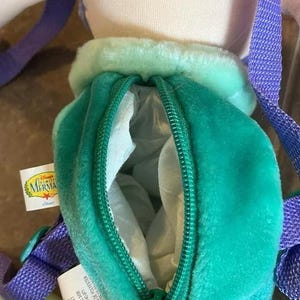 May include: A green and purple plush Ariel backpack with a zippered pocket. The backpack features a Disney The Little Mermaid logo. The tag reads: "Les articles mou Cet article contient du materiel neuf seulement No denreg ont 53 0 3438 No denreg que 1740-871 Contenu fibres de polyester"