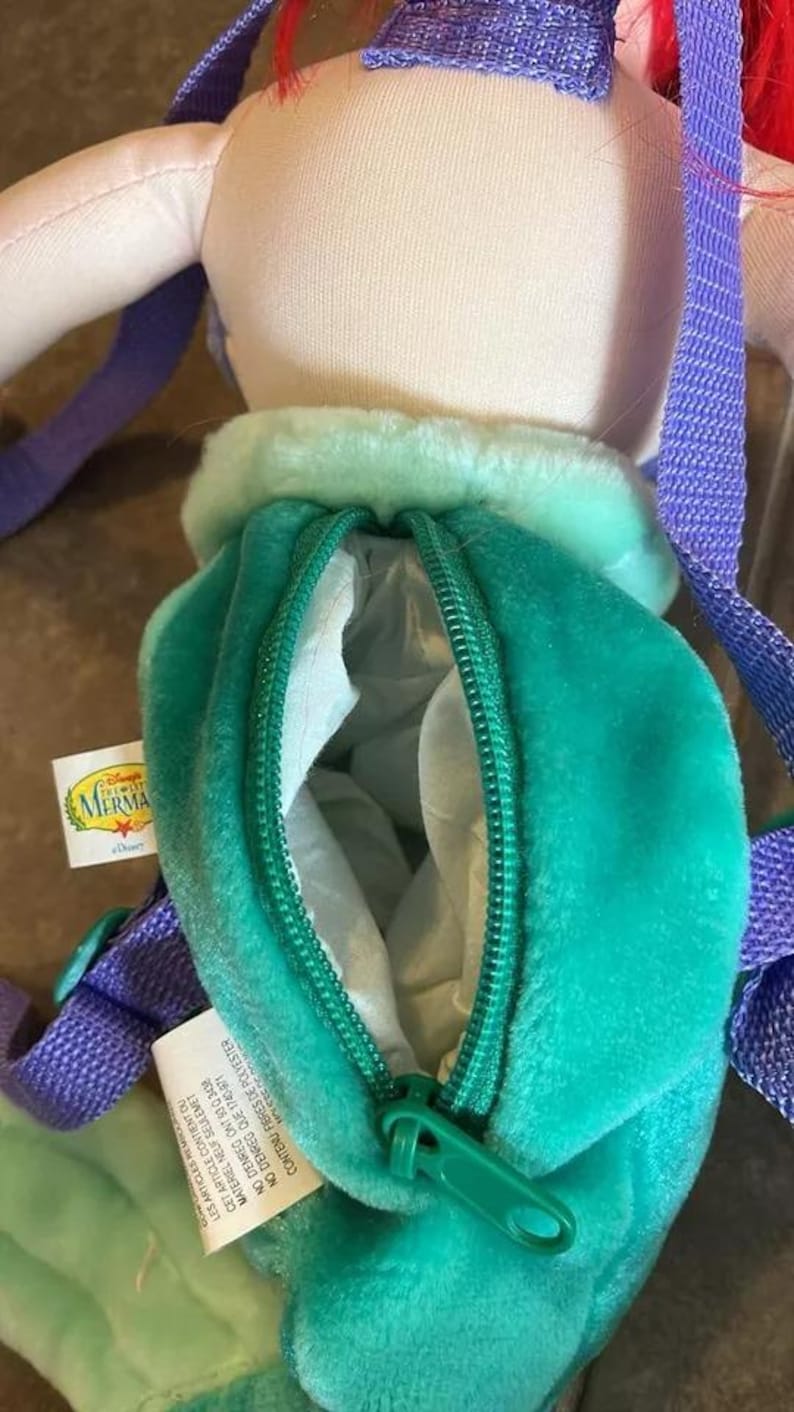 May include: A green and purple plush Ariel backpack with a zippered pocket. The backpack features a Disney The Little Mermaid logo. The tag reads: "Les articles mou Cet article contient du materiel neuf seulement No denreg ont 53 0 3438 No denreg que 1740-871 Contenu fibres de polyester"