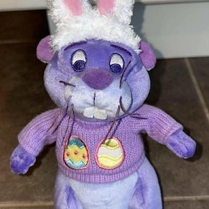 Könnte beinhalten: Ein lila Plüschtier mit einem lavendelfarbenen Pullover mit zwei gestickten Ostereiern. Es hat einen weißen und rosa Hasenohrhut und lange Schnurrhaare. Das Spielzeug ist ein skurriles Oster-Thema.