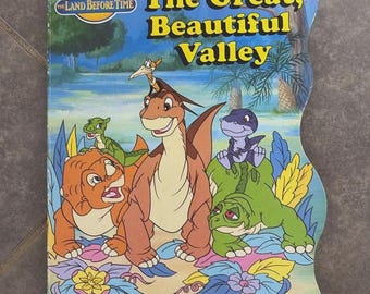 May include: A shaped board book titled The Land Before Time: The Great, Beautiful Valley. The cover features colourful illustrations of cartoon dinosaurs including Littlefoot, Cera, Ducky, Petrie, and Spike in a lush, green prehistoric landscape.