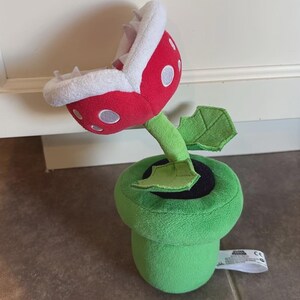 May include: A plush toy featuring a red and white piranha plant with white spots and soft teeth, emerging from a green cylindrical pipe base. The plant has a green stem with two small green leaves attached, and a white tag is visible on the side.