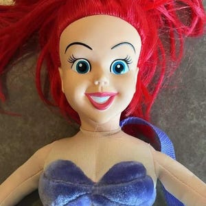 May include: A plush Ariel doll with red hair, a purple seashell bra, and a green tail. The doll is wearing a purple and blue strap.