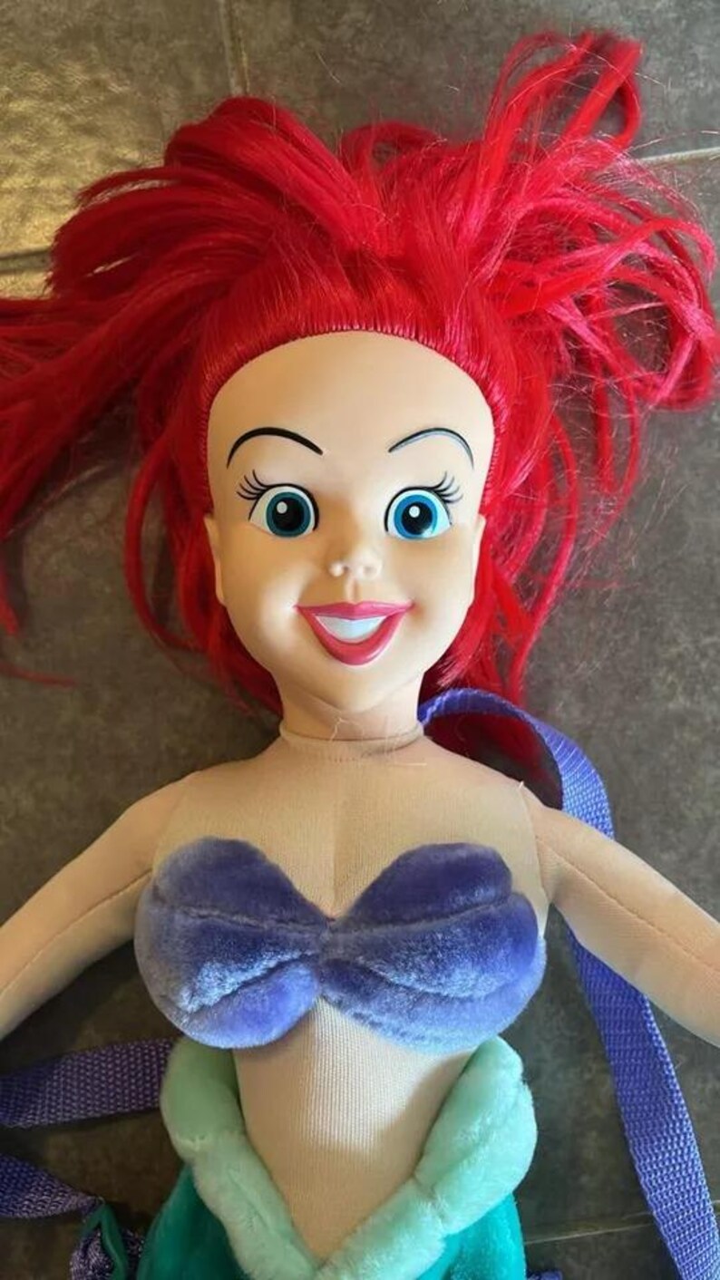 May include: A plush Ariel doll with red hair, a purple seashell bra, and a green tail. The doll is wearing a purple and blue strap.