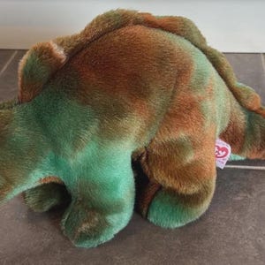 May include: A plush dinosaur toy featuring a soft, fuzzy texture with a mottled pattern of earthy brown and vibrant green colours. The stuffed animal has dark, round plastic eyes and a small, pink Ty brand tag attached to its side near the tail.