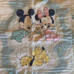 Puede incluir: Una manta de beb&eacute; acolchada blanca con una ilustraci&oacute;n impresa de Mickey Mouse en pijama verde y Minnie Mouse en pijama rosa sentados con un oso de peluche marr&oacute;n. Debajo hay un perro amarillo. La manta est&aacute; decorada con peque&ntilde;as estrellas de colores pastel.