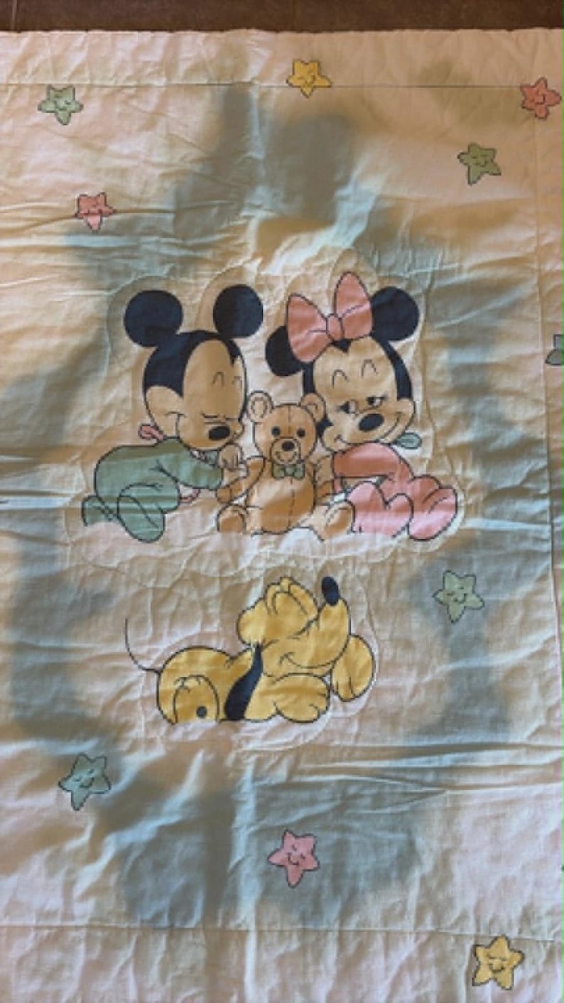 Puede incluir: Una manta de beb&eacute; acolchada blanca con una ilustraci&oacute;n impresa de Mickey Mouse en pijama verde y Minnie Mouse en pijama rosa sentados con un oso de peluche marr&oacute;n. Debajo hay un perro amarillo. La manta est&aacute; decorada con peque&ntilde;as estrellas de colores pastel.