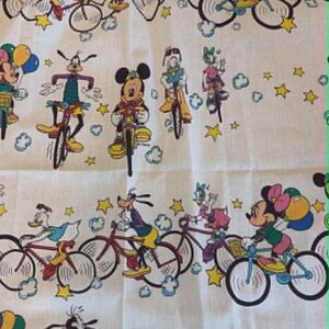 May include: White fabric featuring a repeating pattern of Disney characters riding bicycles. Mickey Mouse, Minnie Mouse, Donald Duck, and Goofy are among the characters depicted. The bicycles are in various colors, and the background includes stars and clouds.