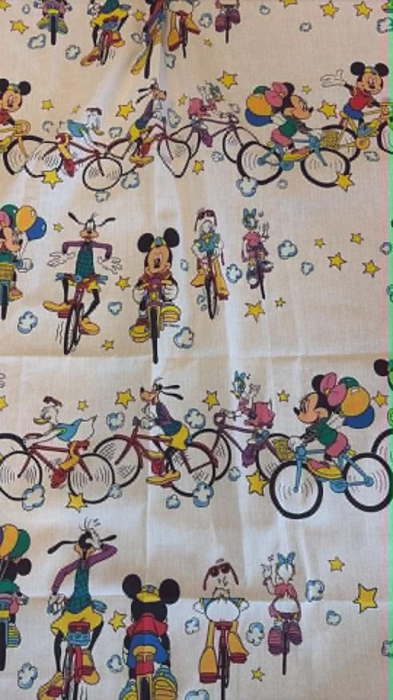 May include: White fabric featuring a repeating pattern of Disney characters riding bicycles. Mickey Mouse, Minnie Mouse, Donald Duck, and Goofy are among the characters depicted. The bicycles are in various colors, and the background includes stars and clouds.
