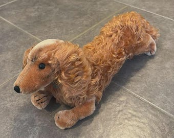Könnte beinhalten: Ein braunes Dackel-Plüschtier mit langem Körper und Schlappohren. Das Spielzeug hat schwarze Augen, eine schwarze Nase und detaillierte Pfoten. Der Hund liegt auf einer grauen Fliesenoberfläche.