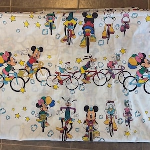 May include: White fabric featuring a repeating pattern of Disney characters riding bicycles. Mickey Mouse, Minnie Mouse, Donald Duck, and Goofy are depicted in colorful outfits, along with yellow stars and clouds. The fabric is likely for bedding or a similar textile.