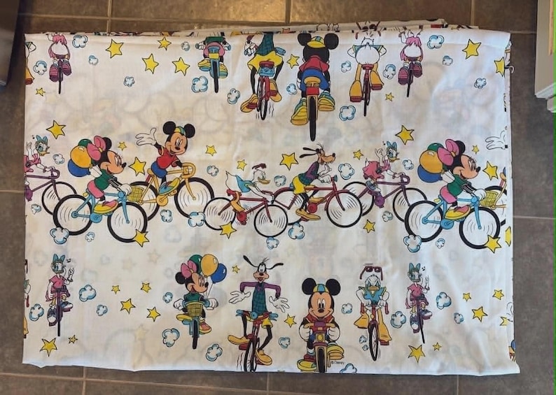 May include: White fabric featuring a repeating pattern of Disney characters riding bicycles. Mickey Mouse, Minnie Mouse, Donald Duck, and Goofy are depicted in colorful outfits, along with yellow stars and clouds. The fabric is likely for bedding or a similar textile.