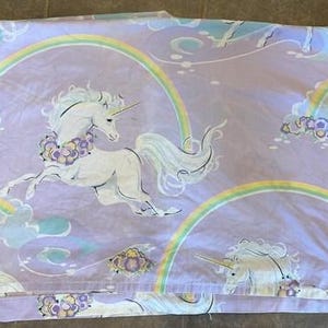 Peut inclure: Un ensemble de draps de couleur lavande avec un motif répété de licornes blanches, d'arcs-en-ciel et de nuages. Les licornes ont des crinières fluides et des guirlandes florales. Les arcs-en-ciel sont multicolores et les nuages sont bleu clair.
