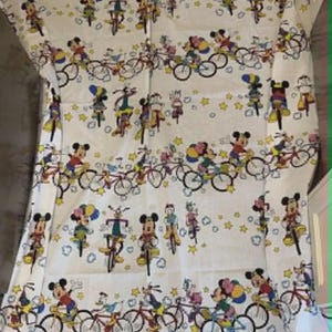 May include: White fabric featuring a repeating pattern of Mickey Mouse and friends riding bicycles. The design includes various characters in colorful outfits, bicycles, and yellow stars. The overall theme is playful and suitable for children's items.