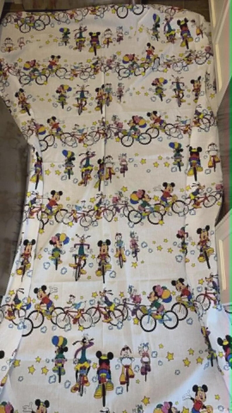 May include: White fabric featuring a repeating pattern of Mickey Mouse and friends riding bicycles. The design includes various characters in colorful outfits, bicycles, and yellow stars. The overall theme is playful and suitable for children's items.