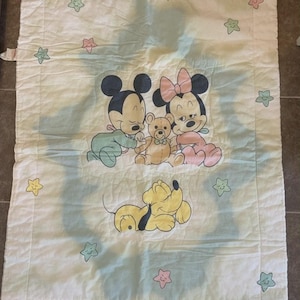 Puede incluir: Una manta rectangular blanca para beb&eacute; con una ilustraci&oacute;n central de los beb&eacute;s Mickey y Minnie Mouse abrazando un oso de peluche, con Pluto durmiendo debajo. El borde est&aacute; decorado con peque&ntilde;as estrellas de colores pastel.